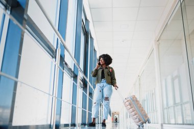 Bavul ve cep telefonuyla yürüyen mutlu bir kadın yolcunun tüm vücut profili.