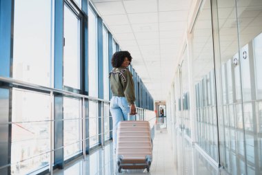 Gündelik giysiler içindeki genç Afro-Amerikan bayan yolcusu havaalanında bavullarıyla bekliyor. Telefonla. Seyahat kavramı.