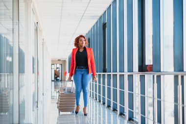 Havaalanında bavuluyla yürüyen siyahi bir kadının tam boy portresi.