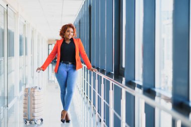 Havaalanında bavuluyla yürüyen siyahi bir kadının tam boy portresi.