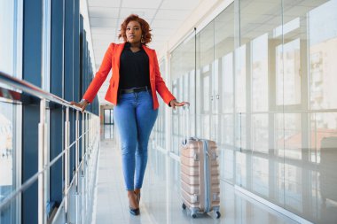 Havaalanında bavuluyla yürüyen siyahi bir kadının tam boy portresi.