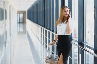 Havaalanında bavuluyla akıllı telefondan konuşan çekici genç bir iş kadınının portresi.
