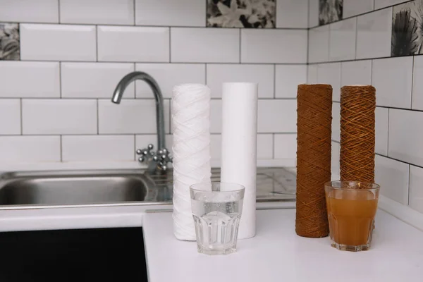 Kullanılan su filtresi kartuşu ve bir bardak paslı su kahverengi boya ve yeni filtre ve bir bardak temiz su. Musluk suyunun demir parçalarıyla kirlendiğine dair kanıt. beyaz mutfak