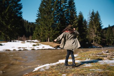 Şapkalı ve yeşil ceketli kadın dağ nehrinin yanındaki dağlarda dinleniyor. Tatil konsepti seyahatinde dinlenme zamanı