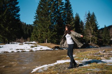 Şapkalı ve yeşil ceketli kadın dağ nehrinin yanındaki dağlarda dinleniyor. Tatil konsepti seyahatinde dinlenme zamanı