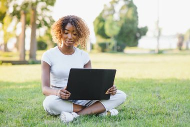 İnsan ve teknoloji kavramı. Kısa peri saç stili çekici Afrikalı kadın güneşli havanın tadını çıkarıyor bahçedeki dizüstü bilgisayarın önünde oturmuş arkadaşını bekliyor.