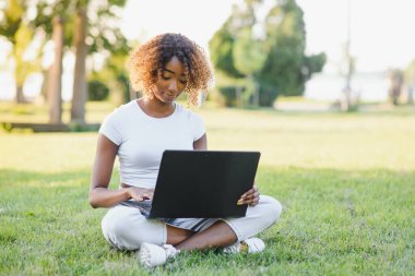Genç, koyu tenli, kolsuz bluz ve kot pantolon giyen çekici bir üniversite öğrencisi güneşli bir günde kampüste oturmuş, tezi üzerinde çalışıyor dizüstü bilgisayar kullanıyor, meşgul ve konsantre görünüyor.