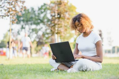 Genç gülümseyen siyah kadın dışarıda çimlerde dizüstü bilgisayarla oturuyor, daktilo yazıyor, internette sörf yapıyor. Teknoloji, iletişim, eğitim ve uzaktan çalışma konsepti, kopyalama alanı