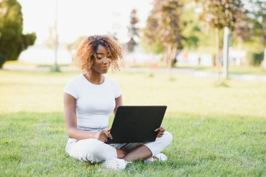 Kıvırcık saçlı, düşünceli, sevimli uluslararası bayan öğrenci halka açık bir parkta, taze çimlerin üzerinde oturuyor, elma ağacına yaslanıyor ve molasında üzüntülü bir şekilde yan tarafa bakıyor.