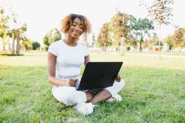 İnsan ve teknoloji kavramı. Kısa peri saç stili çekici Afrikalı kadın güneşli havanın tadını çıkarıyor bahçedeki dizüstü bilgisayarın önünde oturmuş arkadaşını bekliyor.