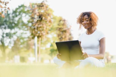 Genç, koyu tenli, kolsuz bluz ve kot pantolon giyen çekici bir üniversite öğrencisi güneşli bir günde kampüste oturmuş, tezi üzerinde çalışıyor dizüstü bilgisayar kullanıyor, meşgul ve konsantre görünüyor.