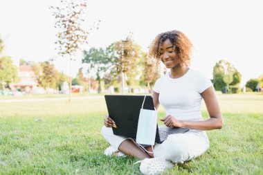 Kıvırcık saçlı, düşünceli, sevimli uluslararası bayan öğrenci halka açık bir parkta, taze çimlerin üzerinde oturuyor, elma ağacına yaslanıyor ve molasında üzüntülü bir şekilde yan tarafa bakıyor.