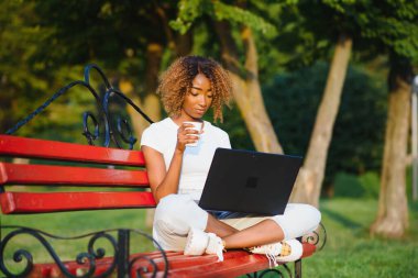 Güzel gülümseyen genç Afrikalı bir kadın dışarıda bir bankta oturuyor, dizüstü bilgisayarla çalışıyor.