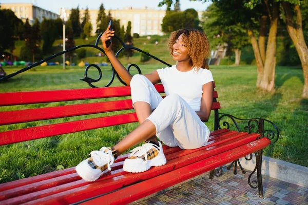 Yeşil pantolonlu, siyah şapkalı, parktaki bankta poz veren ve cep telefonuyla selfie çeken Afro-Amerikan manken kadın..