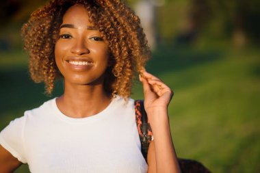 Gün batımında gülümseyen ve parka bakan güzel Afro-Amerikan kadın portresi. Gülümseyen siyah bir kızın açık hava portresi. Neşeli kız, renkli saç bandıyla parkta gülüyor..