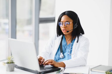 Gülümseyen Afro-Amerikan kadın doktor iş yerinde dizüstü bilgisayar kullanarak beyaz önlük giyiyor, bilgisayarla uzaktan danışmanlık yapıyor, bilgisayar üzerinde çalışıyor, internet teletıp sohbetinde hastaya danışmanlık yapıyor..