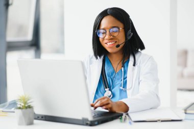 Gülümseyen Afro-Amerikan kadın doktor iş yerinde dizüstü bilgisayar kullanarak beyaz önlük giyiyor, bilgisayarla uzaktan danışmanlık yapıyor, bilgisayar üzerinde çalışıyor, internet teletıp sohbetinde hastaya danışmanlık yapıyor..