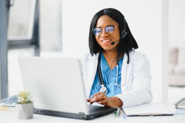 Afrikalı doktor kulaklık takıyor hastaya danışmanlık yapıyor. Bilgisayarda web kamerasından görüntülü arama yapıyor. Teletıp video konferansı uzaktan bilgisayar uygulaması sanal toplantısı. Omuz üzerinden video çağrısı görünümü.