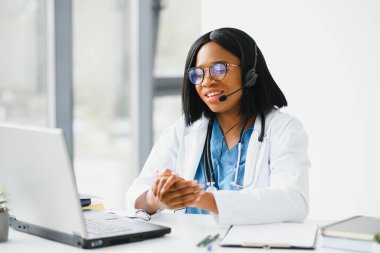 Gülümseyen Afro-Amerikan kadın doktor iş yerinde dizüstü bilgisayar kullanarak beyaz önlük giyiyor, bilgisayarla uzaktan danışmanlık yapıyor, bilgisayar üzerinde çalışıyor, internet teletıp sohbetinde hastaya danışmanlık yapıyor..