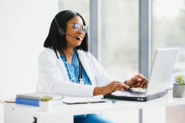 Afrikalı doktor kulaklık takıyor. Siyahi kadın hastaya danışıyor. Bilgisayarda web kamerasından görüntülü arama yapıyor. Teletıp video konferansı uzaktan bilgisayar uygulaması sanal toplantısı. Omuz üzerinden video çağrısı görünümü.