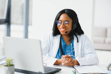 Afrikalı doktor kulaklık takıyor hastaya danışmanlık yapıyor. Bilgisayarda web kamerasından görüntülü arama yapıyor. Teletıp video konferansı uzaktan bilgisayar uygulaması sanal toplantısı. Omuz üzerinden video çağrısı görünümü.