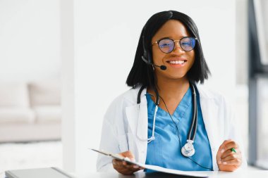 Gülümseyen Afro-Amerikan kadın doktor iş yerinde dizüstü bilgisayar kullanarak beyaz önlük giyiyor, bilgisayarla uzaktan danışmanlık yapıyor, bilgisayar üzerinde çalışıyor, internet teletıp sohbetinde hastaya danışmanlık yapıyor..
