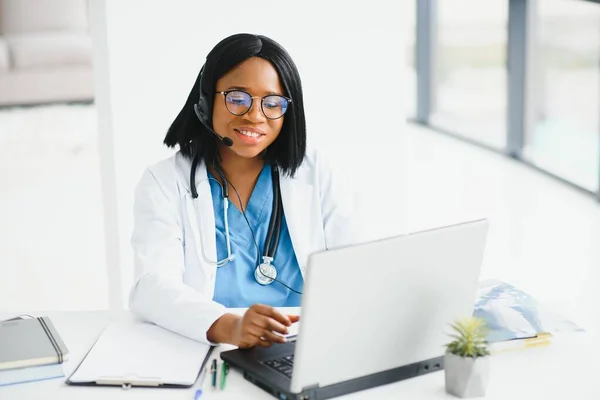 Gülümseyen Afro-Amerikan kadın doktor iş yerinde dizüstü bilgisayar kullanarak beyaz önlük giyiyor, bilgisayarla uzaktan danışmanlık yapıyor, bilgisayar üzerinde çalışıyor, internet teletıp sohbetinde hastaya danışmanlık yapıyor..