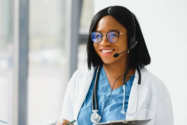 Smiling african american female doctor gp wears white medical coat using laptop computer at workplace gives remote online consultation, working on pc, consulting patient in internet telemedicine chat.