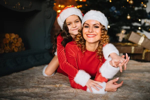 Beautiful young mother and daughter sitting on the floor next to a Christmas tree, hugging.