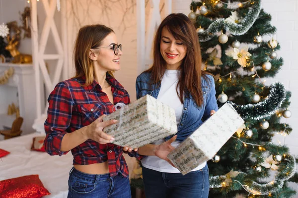 Two girlfriends give gift boxes to each other near a Christmas tree.