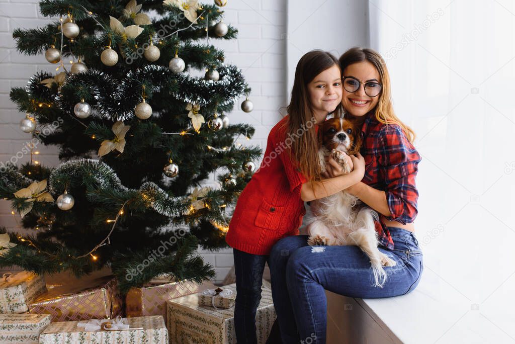 Happy winter holidays! Positive brunette woman embraces little girl pose with gifts on floor in room, dog near, have fun near Christmas tree.
