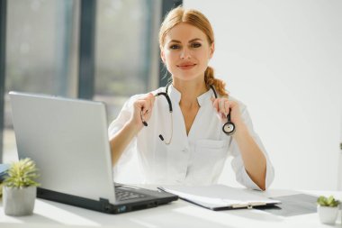 Ofis masasında çalışan ve kameraya gülümseyen kadın doktor, arka planda ofis içi.