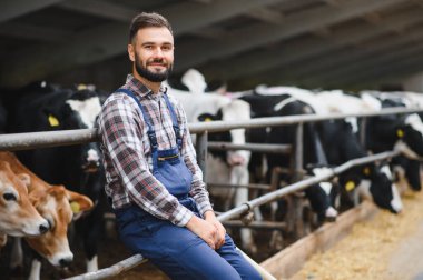 Arka planda ineklerle çevrili bir ahırda oturan ve gülümseyen genç bir çiftçinin portresi, neşeli bir kırsal yaşam tarzını yansıtıyor.