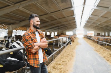 Genç çiftçi ahırda durup saman yerken kollarını kavuşturup gülümsüyor.