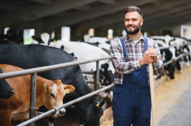 Kolları çapraz duran, elinde bir dirgenle ahırda ineklerle gülümseyen genç bir çiftçinin portresi.