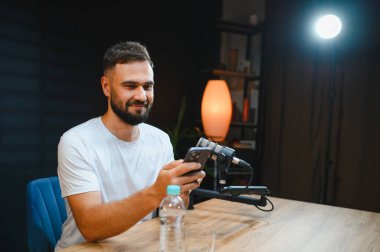 Genç adam akıllı telefon ve mikrofon kullanarak ses içeriğini kaydediyor. Hafif lamba ışığıyla aydınlatılıyor.