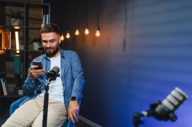 Akıllı telefon ve mikrofon kullanan içerik yaratıcısı, sıcak ışıkları ve mavi arka planı olan ev stüdyolarında podcast yapıyor.