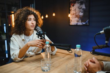 İki podcaster, bir erkek ve bir kadın, profesyonel bir stüdyoda ses içeriğini kaydederken dinamik sohbetler yapıyorlar.