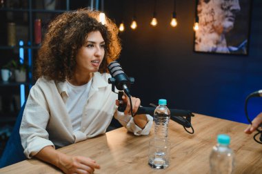 Gelişmiş ses ekipmanlarıyla çevrili modern bir profesyonel stüdyoda podcast yaparken mikrofona konuşan genç bir kadın.