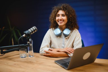 Çevrimiçi içerik oluşturan genç bir kadın, profesyonel mikrofon ve dizüstü bilgisayar kullanarak bir ev stüdyo ortamında bir vlog veya podcast kaydediyor.