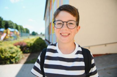 Gözlüklü ve sırt çantalı neşeli bir öğrencinin portresi renkli okul binasının önünde gülümseyerek öğrenmeye ve çalışmaya hazır.