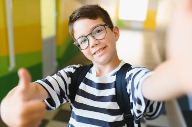 Sırt çantalı, gözlüklü, selfie çeken ve okul koridorunda baş parmak hareketi yapan mutlu çocuk.