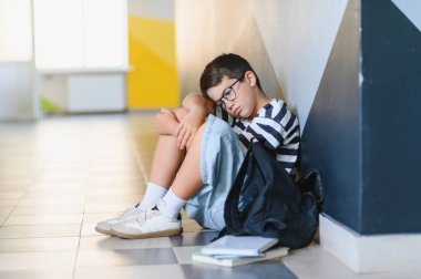 Üzgün okul çocuğu okulun koridorunda tek başına oturmuş, zorbalıktan acı çektikten sonra dizlerine sarılıp ağlıyor.