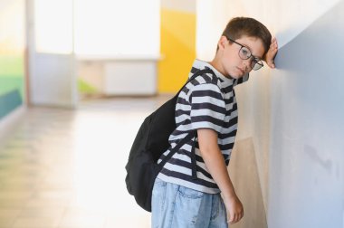 Okul koridorunda duvara yaslanmış sırt çantalı bir öğrenci, sınıf arkadaşlarının zorbalığının ağırlığını hissediyor ve üzüntü yaşıyor.