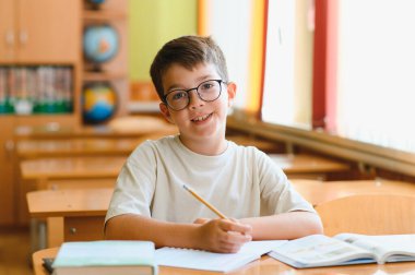 İlkokul öğrencisi deftere yazı yazıyor, okul masasında çalışıyor, gülümsüyor ve kameraya bakıyor.