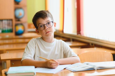 İlköğretim öğrencisi bir masada çalışırken özenle bir not defterine yazıyor, hareketli bir sınıfın içinde öğrenmeye dalmış.