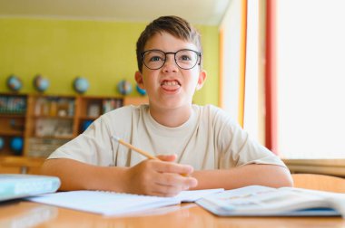 Genç bir çocuk elinde kalem tutarken komik bir yüz ifadesi takınıyor. Canlı bir sınıfın ortasında masa başında ders çalışıyor.