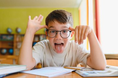 Gözlüklü genç bir çocuk komik bir yüz ifadesi takınıyor ve sınıftaki bir masada otururken dilini çıkarıyor.