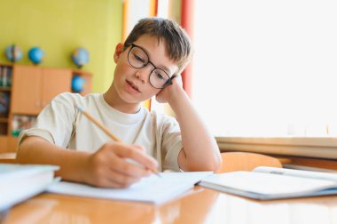 Genç öğrenci gayretli bir şekilde bir not defterine yazıyor, hareketli bir sınıftaki masada otururken derslerine odaklanıyor.