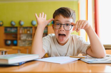 Gözlüklü öğrenci komik bir yüz ifadesi takınıyor ve sınıfta masa başında otururken dilini dışarı çıkarıyor. Öğrenmeye karşı neşeli bir tavır sergiliyor.